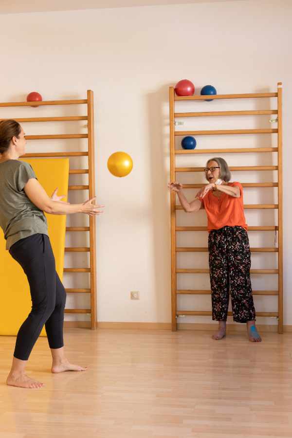 Frau Olescher behandelt im großen Gymnastikraum eine ältere Dame indem sie ihr einen gelben Ball zuwirft.